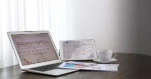 Laptop and tablet on a wooden desk displaying financial charts and data, with documents, a smartphone, and a coffee cup nearby.