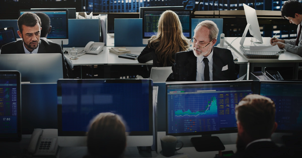 Financial traders at work in a busy office with monitors displaying stock charts and data in a high-rise city setting.
