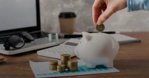 Hand deposits euro coin into white piggy bank on desk with laptop, coins, calculator, and chart; symbolizes saving, finance, and budgeting.