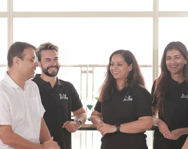 Four smiling adults stand indoors, chatting warmly. Three wear "hola prime" shirts